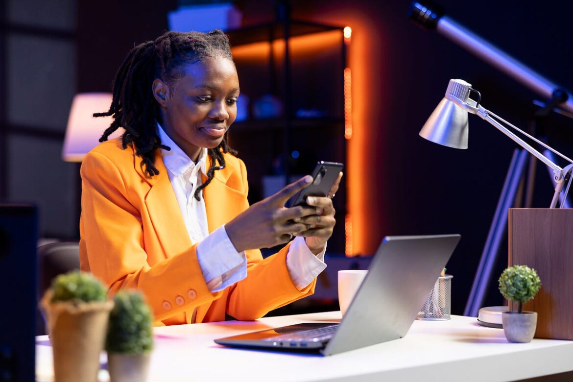 A young Black woman with dreadlocks sits at a desk in a modern, brightly lit home office at night. She is smiling while looking at her smartphone, which she holds with both hands. An open laptop, a white mug, a desk lamp, and small potted plants are visible on the desk in front of her.