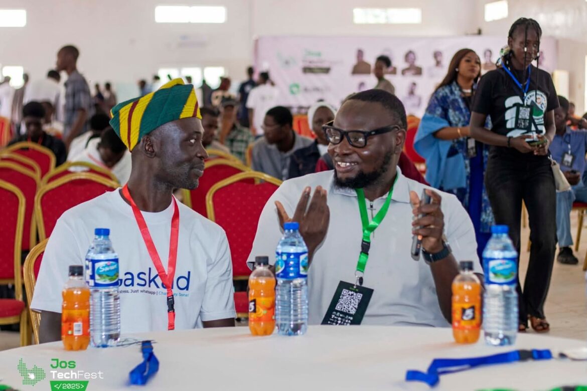 Two men are seated at a table during the Jos TechFest event. The man on the left, wearing a distinctive yellow and green cap and a white t-shirt, is looking at the man on the right. The man on the right, wearing glasses and a light gray shirt with a lanyard, is actively speaking and gesturing with his hands. Several water and orange soda bottles are on the table between them. The background shows other attendees and red chairs.